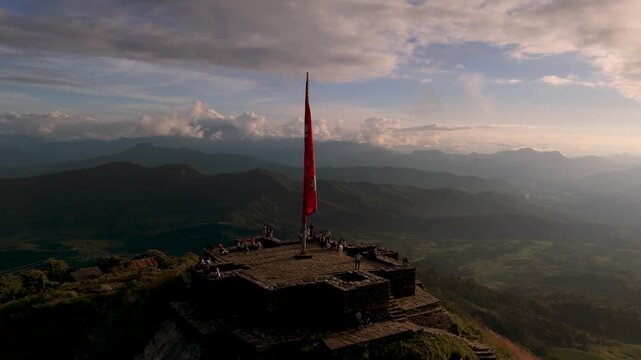 Majestic 4K aerial view of Nepalese flag flying on a stone platform view tower, tourists visiting hilltop destination in the Himalayas, blue sky and green hills background