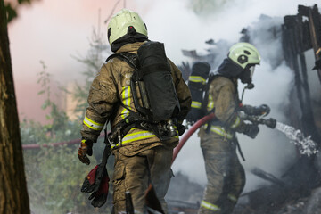 Two professional firefighters in full gear and oxygen tanks battling a blaze, spraying water onto a charred, collapsed structure surrounded by thick smoke.