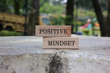 A pair of wooden blocks with the words POSITIVE and MINDSET sit stacked on a weathered stone ledge in a sunny park. conveys motivation, optimism, and a mindful, growth oriented attitude.