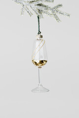 Minimal creative view of Glass of white wine Christmas decoration hang on snow green fir branch. New Year glass toy as wineglass with gold glitters. Festive minimal concept xmas party, winter holiday.