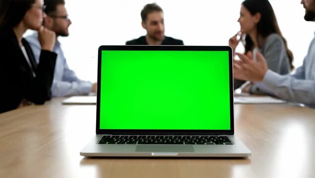 Laptop with a green screen for mockup on a conference table during a corporate business meeting with a team of colleagues in the background