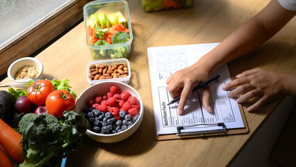 Nutritionist analyzing and writing a healthy meal plan with fresh fruits, vegetables, and nuts on...