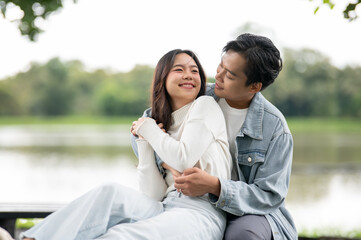 Obraz premium Young asian couple woman getting back hug or waist holding by man sitting outdoors on bench in park.