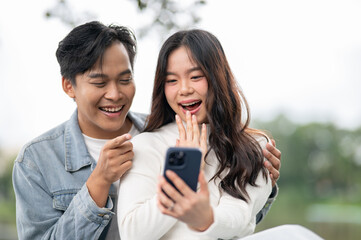 Fototapeta premium Young asian couple looking at phone a man hugging woman's shoulder while sitting outdoors in park.