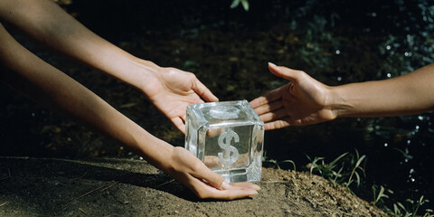 Two hands exchanging transparent glass cube with dollar sign symbolizing financial flexibility and trust in natural outdoor setting