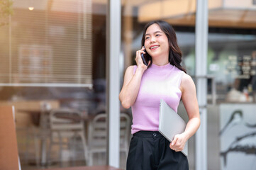 Fototapeta premium Young asian woman talking on phone as carrying a laptop while standing or walking in front of a cafe