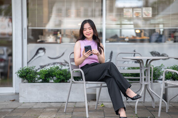 Young asian woman holding and looking at phone with smile while sitting at table in front of a cafe