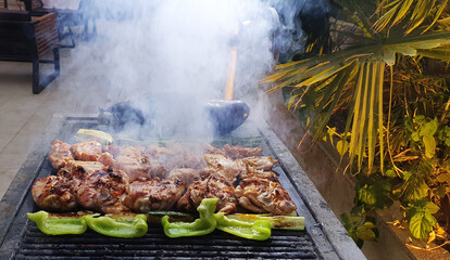 Grilled chicken pieces and vegetables sizzling on an outdoor barbecue grill surrounded by rising smoke and garden greenery.