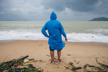 back of man tourist in a raincoat on a dirty beach by the sea after a storm in cloudy weather. Concept of the rainy and monsoon season