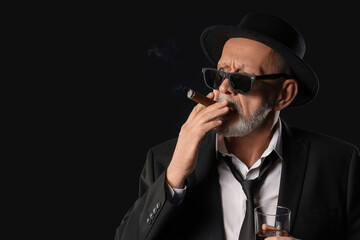 Old businessman with glass of whiskey smoking cigar on black background