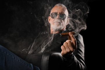 Stylish old man in leather jacket smoking cigar on black background