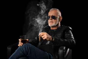 Stylish old man in leather jacket with glass of whiskey smoking cigar on black background