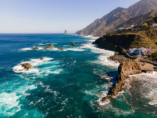 High angle aerial view of the rugged northern shoreline of Tenerife, Spain, with striking azure blue sea, dark volcanic rocks, and foamy waves creating vivid contrast.

