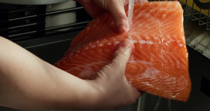 Anonymous female is preparing for lunch at home. The housewife washes salmon. Crop woman hand washing fresh raw salmon slices under running water at kitchen sink.