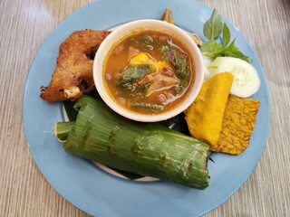 Traditional Indonesian lunch menu consists of sayur asam (or sayur asem) in a bowl, fried chicken, rice wrapped in banana leaves, tempeh, and tofu. Sayur asam is made from tamarind, it tastes sour