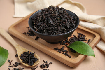 Bowl with dry black tea, leaves and spoon on beige background