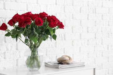 Vase with beautiful red roses, magazines and decor on chest of drawers near white wall, closeup