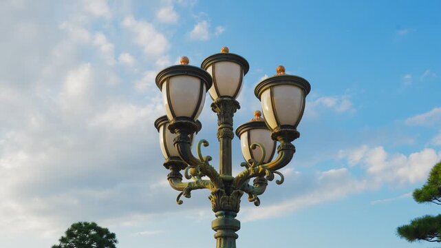 A low-angle close-up shot of a decorative, multi-headed vintage lamppost with ornate metalwork set against a bright, partly cloudy blue sky.