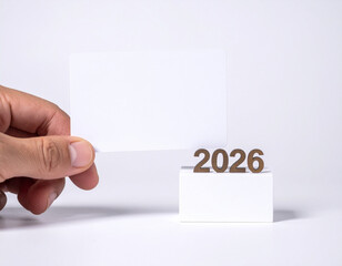 Obraz premium A hand holds a blank card near the year 2026 in wooden numbers, set on a white block against a white background.