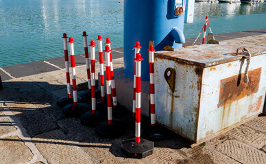 Red and White Striped Traffic Poles Stacked Next to a Rusty Metal Box and Blue Utility Pillar on the Stone Pavement of a Mediterranean Harbor