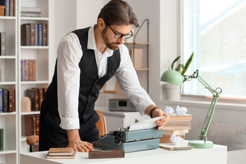 Young male writer with vintage typewriter, crumpled papers and books working in office