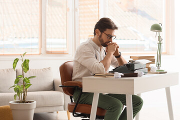 Young male writer with vintage typewriter, crumpled papers and books near window in office