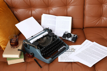 Vintage typewriter, photo camera, pipe and books on brown leather sofa in living room, closeup