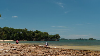 A serene coastal scene with people walking along a rocky beach under clear blue skies. Lush green trees line the shore, and distant islands rest peacefully across calm tropical waters on a sunny day.