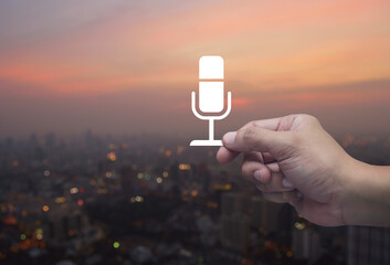 Hand holding microphone flat icon in fingers over blur of cityscape on warm light sundown, Technology communication concept