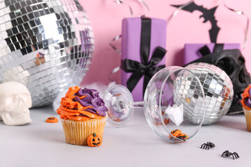 Tasty cupcake with disco balls, skulls, gift boxes, glass and Halloween decor on color background