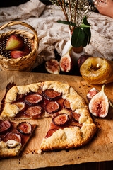 Rustic homemade fig galette on parchment paper. A dark and moody food photography still life with low key lighting, served on a vintage wooden table with a wicker basket and lace.