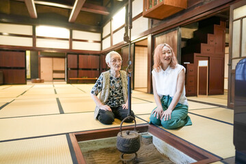 Two women sitting on a tatami mat in a room with a pot on the floor. The room has a traditional...