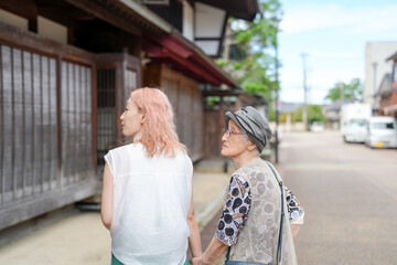 Fototapeta premium Two women walking down a street, one with pink hair. The woman with the pink hair is holding the hand of the older woman