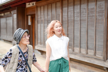 An older woman with gray hair is walking with a younger woman who has red hair. The older woman is wearing a hat and a vest, while the younger woman is wearing a white shirt and green pants