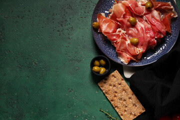 Plate with delicious prosciutto slices, olives and crispbread on green background