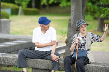 Two older people are sitting on a bench, one of them holding a cane. They seem to be enjoying each other's company and the peaceful atmosphere of the park. The man is wearing a blue hat