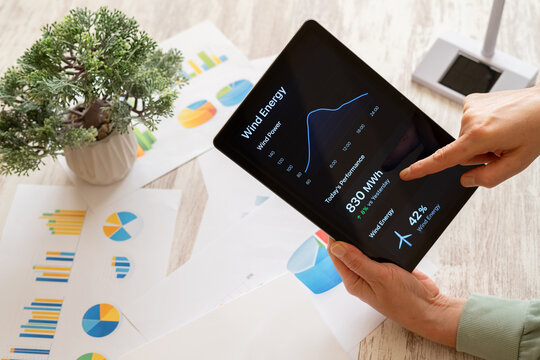 Hands holding a digital tablet tracking wind energy performance data and renewable power statistics on a modern application, surrounded by financial charts and a small plant