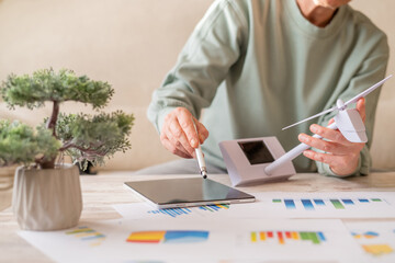 Person holds wind turbine model while tapping tablet with charts and data, visualizing renewable energy research, green technology planning and sustainable power development