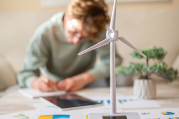 Person studying renewable energy data and making notes next to a wind turbine model, reflecting concepts of sustainable technology, ecological planning, and future smart energy