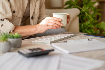 Woman holding a mug while organizing personal finances, calculating expenses, and planning savings from a home office workstation, reflecting modern freelance work and economy