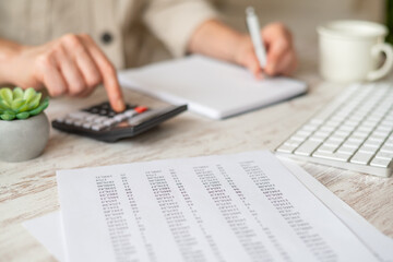 Woman managing personal finances, calculating expenses and income using a calculator, keeping records in a notebook, and organizing financial data on printed reports at a home office desk