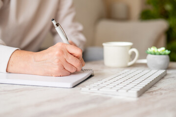 Fototapeta premium Woman's hand holding a pen, writing notes in a spiral notebook, showing ideas of budgeting, personal finance, home economics, and financial planning on a desk with a keyboard and coffee cup