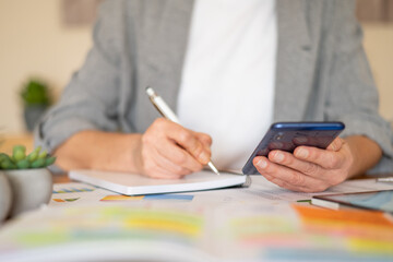 Woman balancing productivity and personal planning, writing in a notebook while checking her smartphone to schedule tasks, manage time and stay organized at her desk