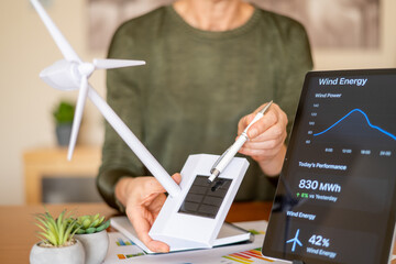 Person holding a model wind turbine with a solar panel, pointing to a tablet showing a wind energy performance application, symbolizing green technology and sustainable future