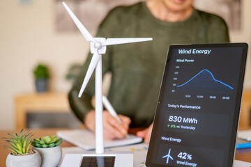 Person analyzing wind energy data on a digital tablet next to a miniature wind turbine model, symbolizing sustainable energy management and ecological future planning