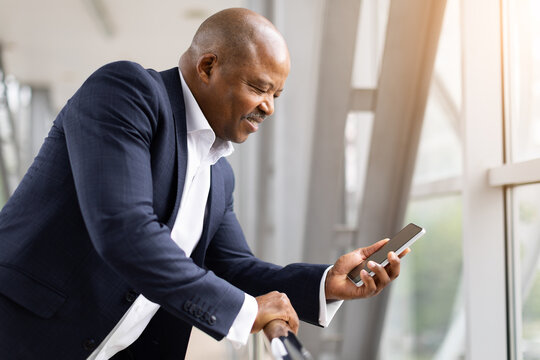 Smiling African American businessman leaning on railing at airport terminal, checking phone and enjoying travel moment, sunlight reflecting on background