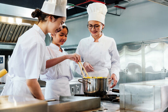 Team of chefs in white uniforms and toques stir pasta on stove during culinary class. Students and instructor show collaboration, education, teamwork, skill, and hands-on lesson in kitchen setting.