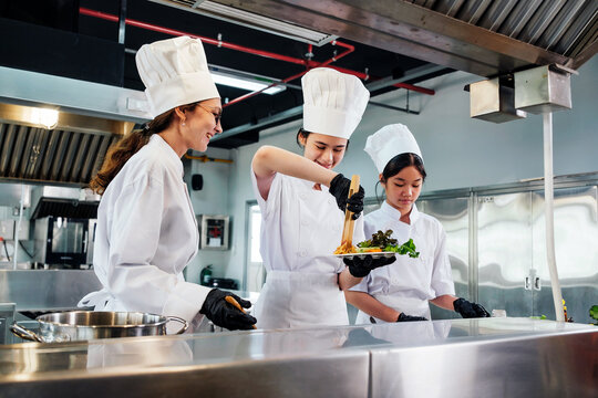 Team of chefs in white uniforms and toques plate gourmet dish in commercial kitchen. Scene shows instructor-led collaboration, dining presentation, stainless workflow, education, teamwork in school. - Powered by Adobe