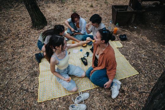 A group of young friends sit on a yellow checkered blanket in a wooded park, sharing food, chatting, and enjoying a casual outdoor picnic together. - Powered by Adobe
