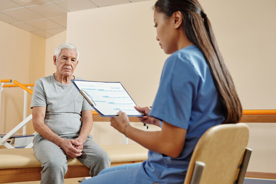 Young professional female physiotherapist looking at medical record of elderly Caucasian male patient sitting on couch in rehab center